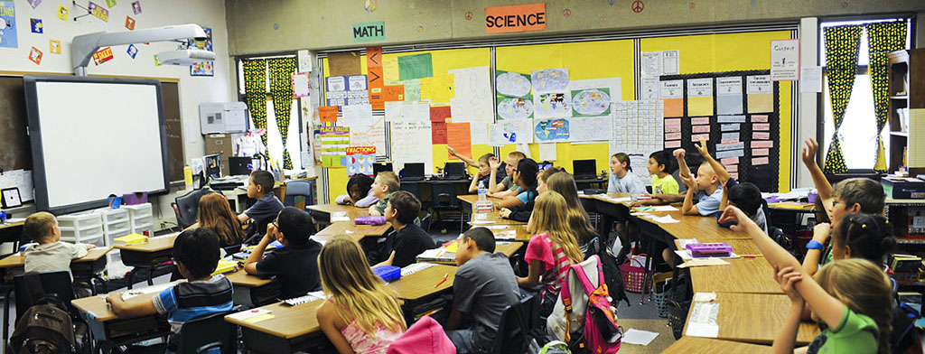 The image is of a class of about 23 elementary students. The rows of desks are arranged in four arching rows, with rows closer to the back having more students. All desks and students are facing the front of the classroom. Six students throughout the back two rows are raising their hands.